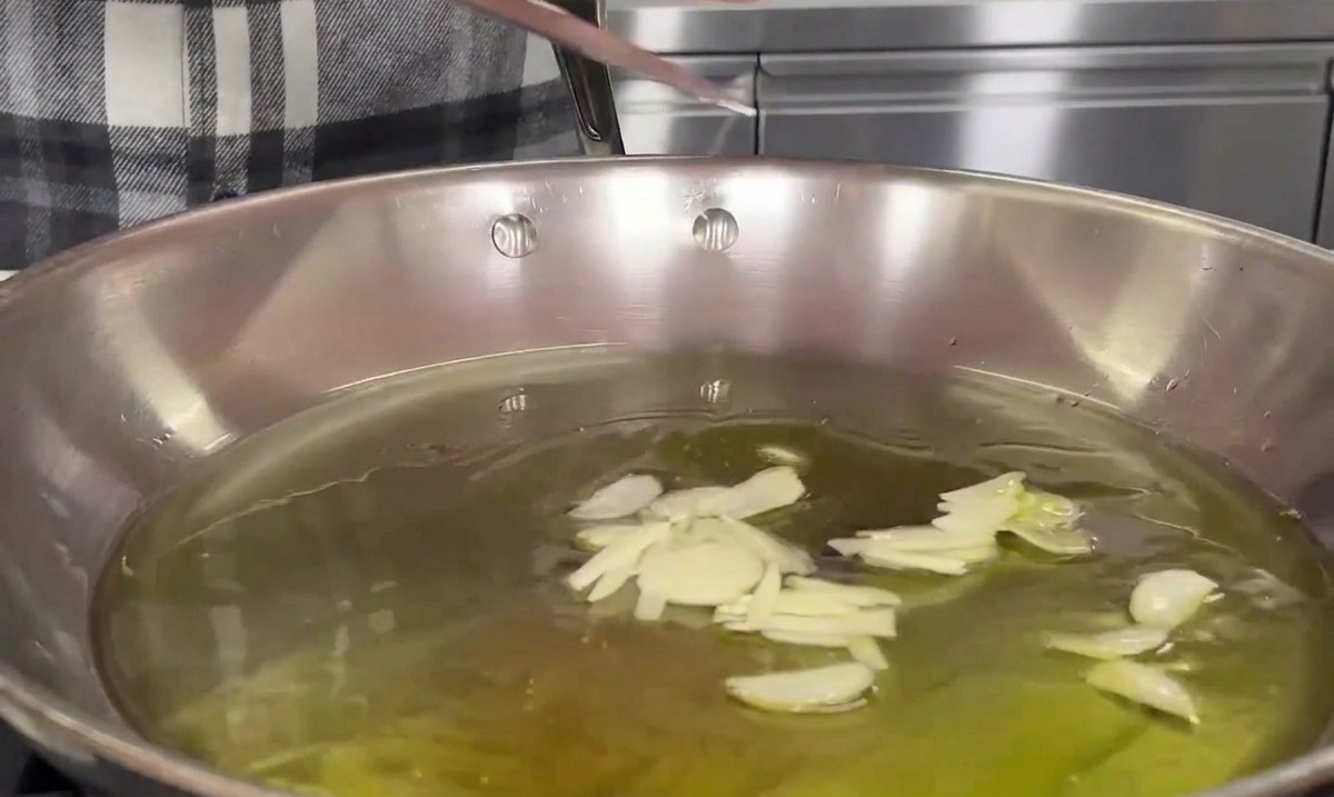Knoblauchscheiben gleiten von einer Messerklinge in eine Pfanne mit warmem Olivenöl.