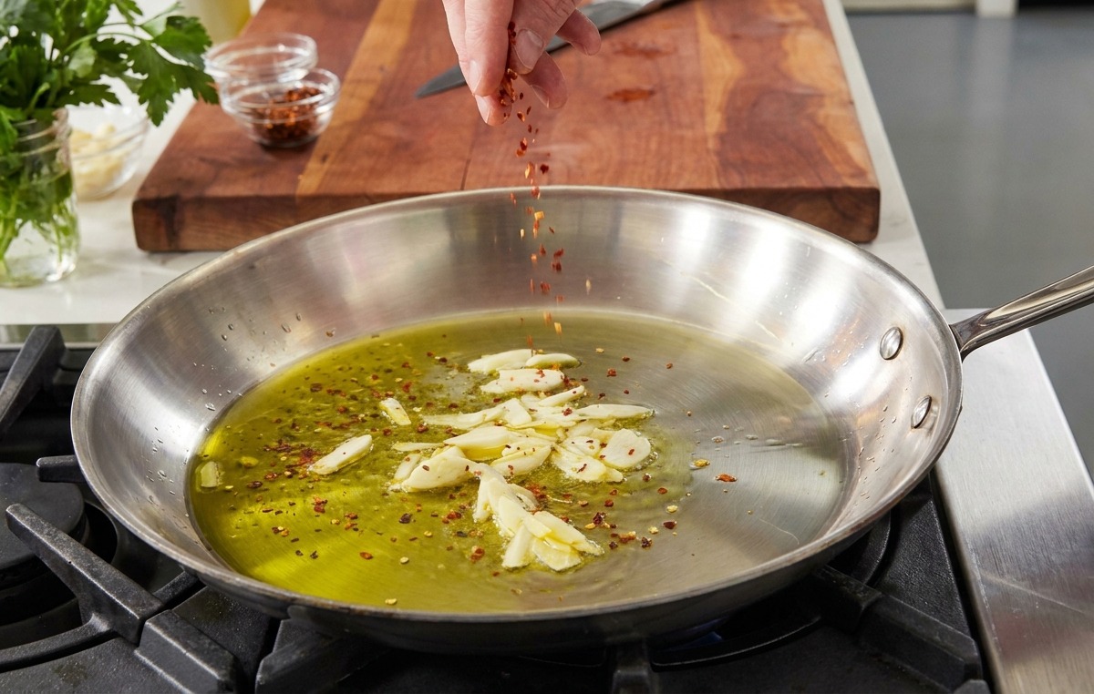 Eine Hand streut rote Chiliflocken in eine Pfanne mit Olivenöl und Knoblauchscheiben.
