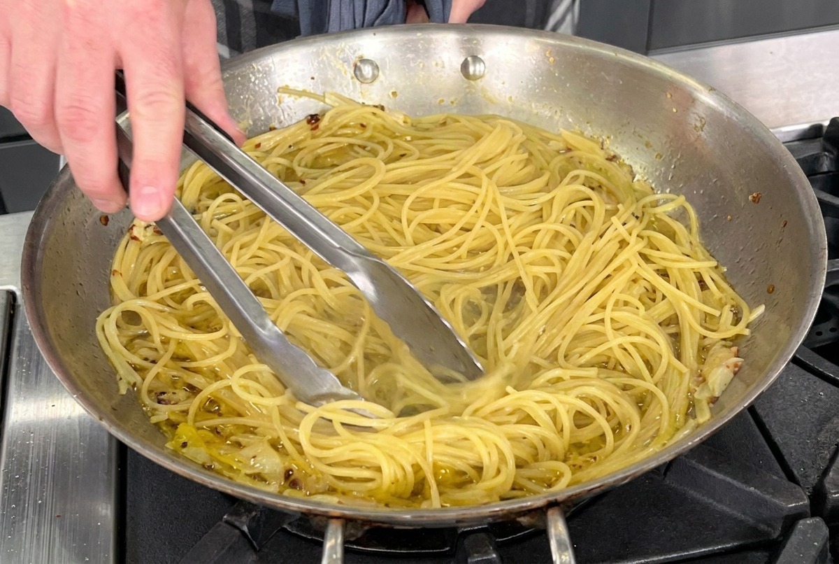 Spaghetti werden mit einer Zange in einer Edelstahlpfanne mit Olivenöl, Knoblauch und Chili geschwenkt.