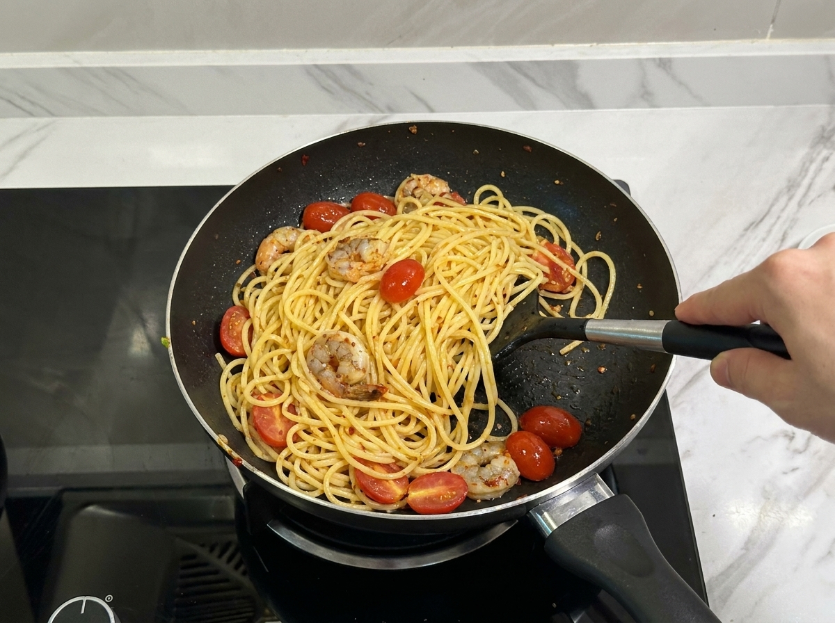 Ein Pfannenwender mischt Spaghetti, Garnelen und Tomaten in der Pfanne.