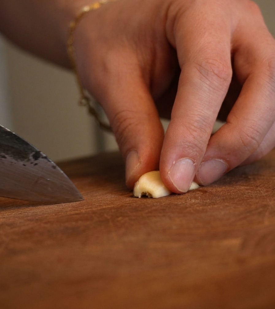 Nahaufnahme von Fingern, die Knoblauch halten, während ein Kochmesser ihn in feine Stücke hackt.