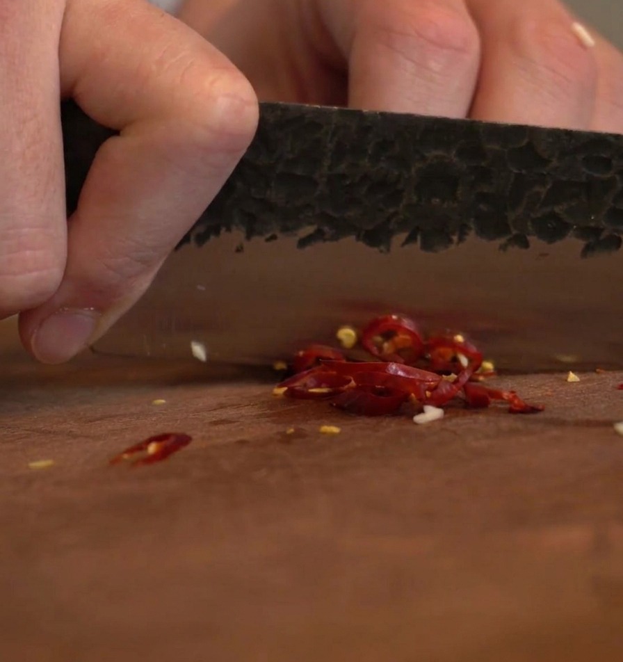 Eine dunkle Messerklinge hackt getrocknete rote Chilischoten in kleine Flocken auf einem Brett.