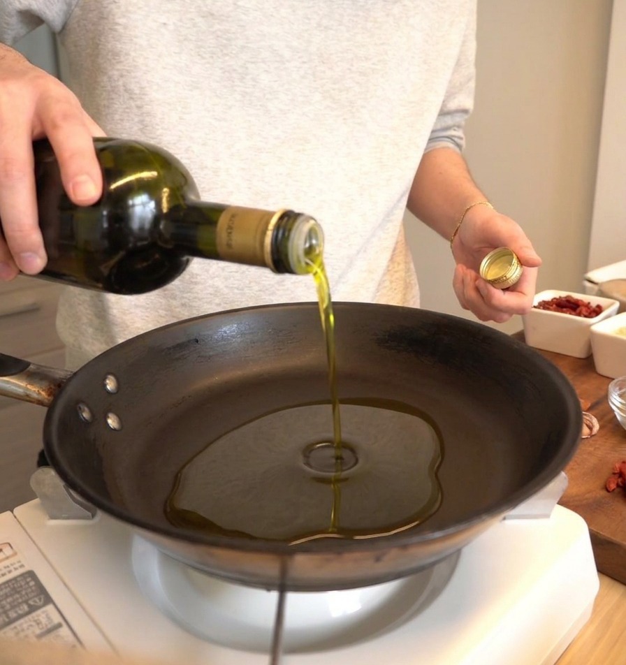 Hochwertiges Olivenöl wird aus einer dunklen Glasflasche in eine kalte schwarze Pfanne gegossen.