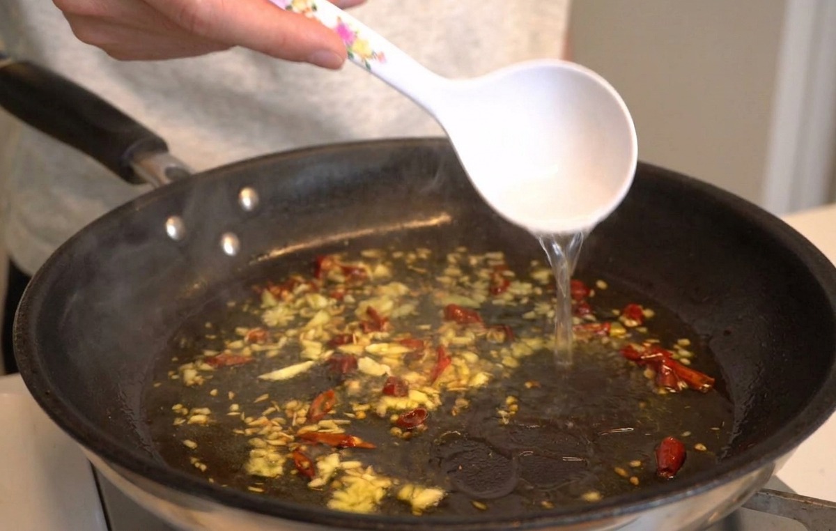 Eine weiße Kelle gießt heißes, trübes Pastawasser in die Pfanne mit Knoblauch, Öl und Chili.