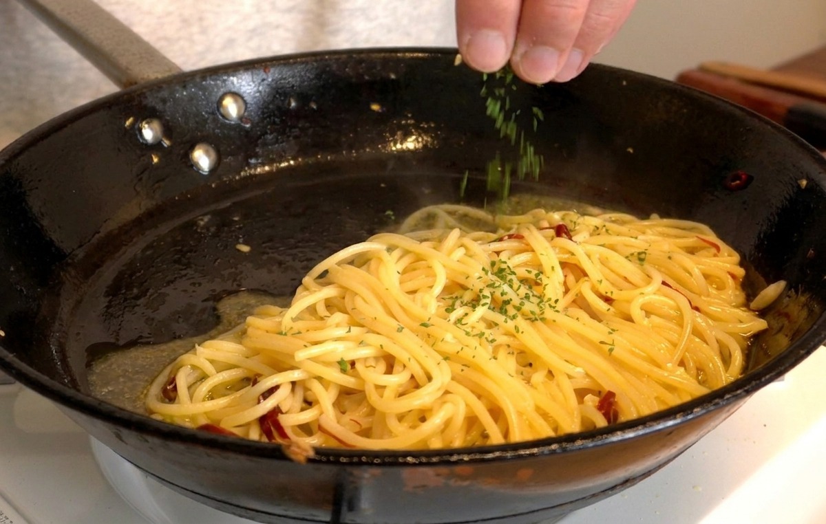 Eine Hand streut frisch gehackte Petersilie über die glänzenden Spaghetti in der Pfanne.