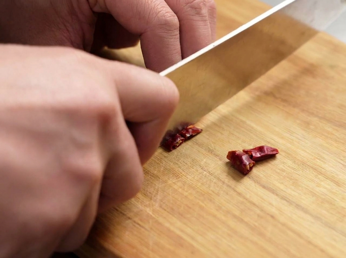 Ein Messer, das getrocknete rote Chilischoten auf einem Holzbrett in kleine Stücke schneidet.