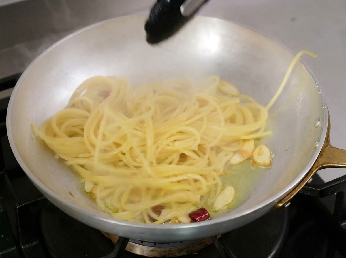 Gekochte Spaghetti, die mit einer schwarzen Zange direkt in eine Pfanne mit heißem Knoblauch-Chili-Öl gehoben werden.