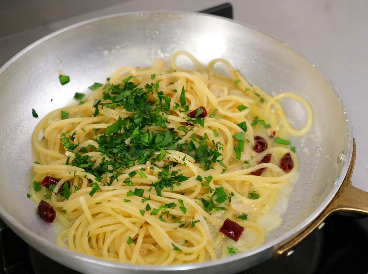 Fein gehackte frische Petersilie, die großzügig über gekochte Spaghetti in einer Metallpfanne gestreut wird.