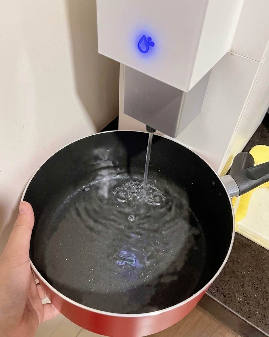Eine Hand hält einen roten Topf unter einen Wasserspender zum Befüllen.