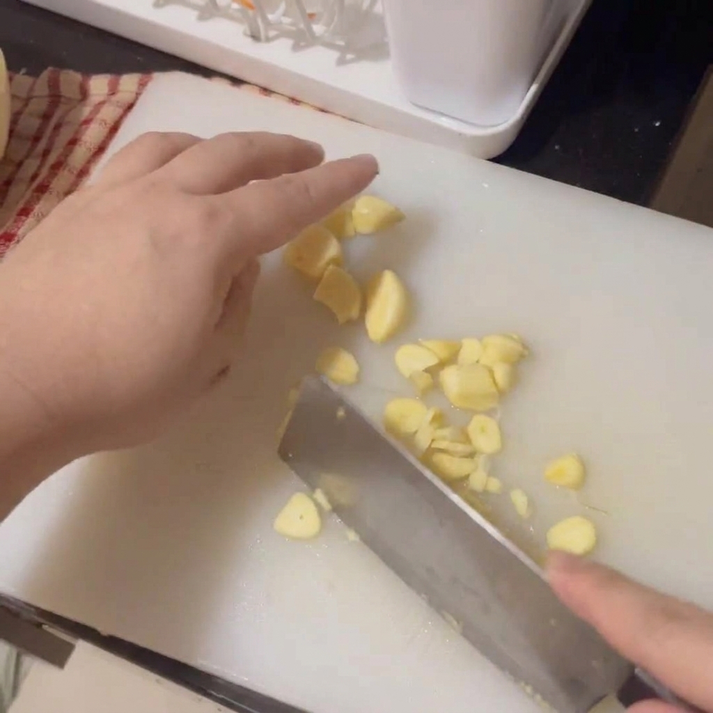 Hände schneiden frische Knoblauchzehen mit einem Hackmesser auf einem weißen Brett in feine Scheiben.