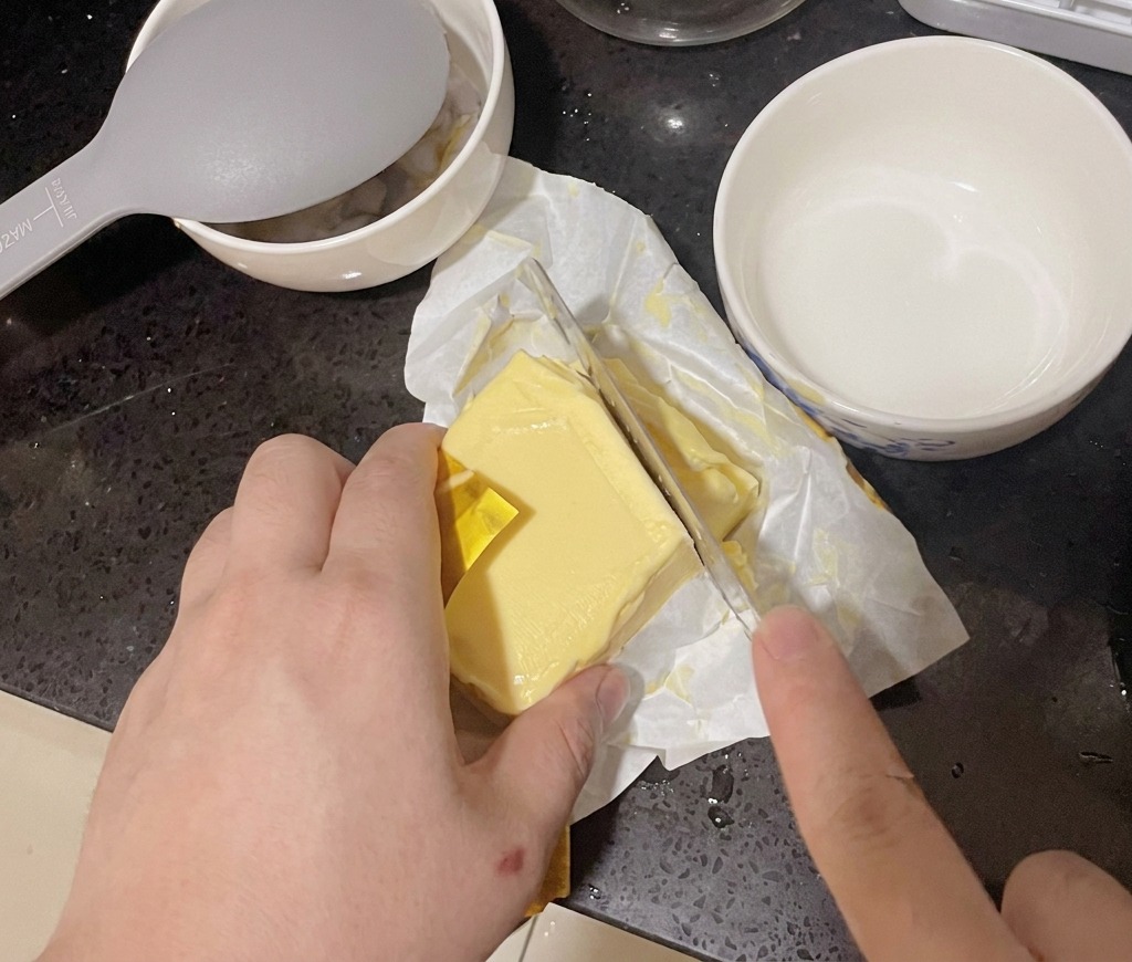 Eine Person schneidet mit einem großen Küchenmesser ein Stück von einem Butterblock ab.