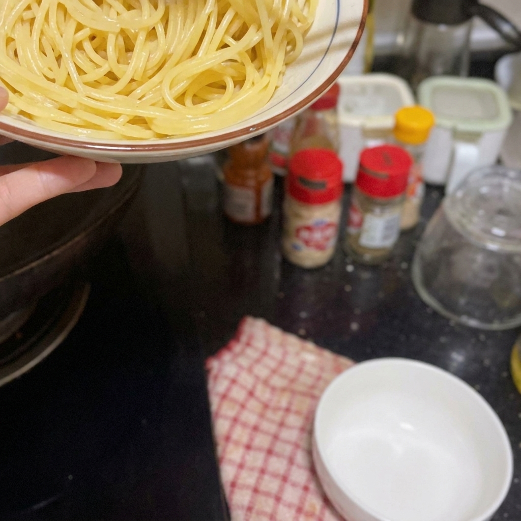 Eine Hand hält eine Schüssel mit Spaghetti über den Herd, um sie in die Pfanne zu geben.