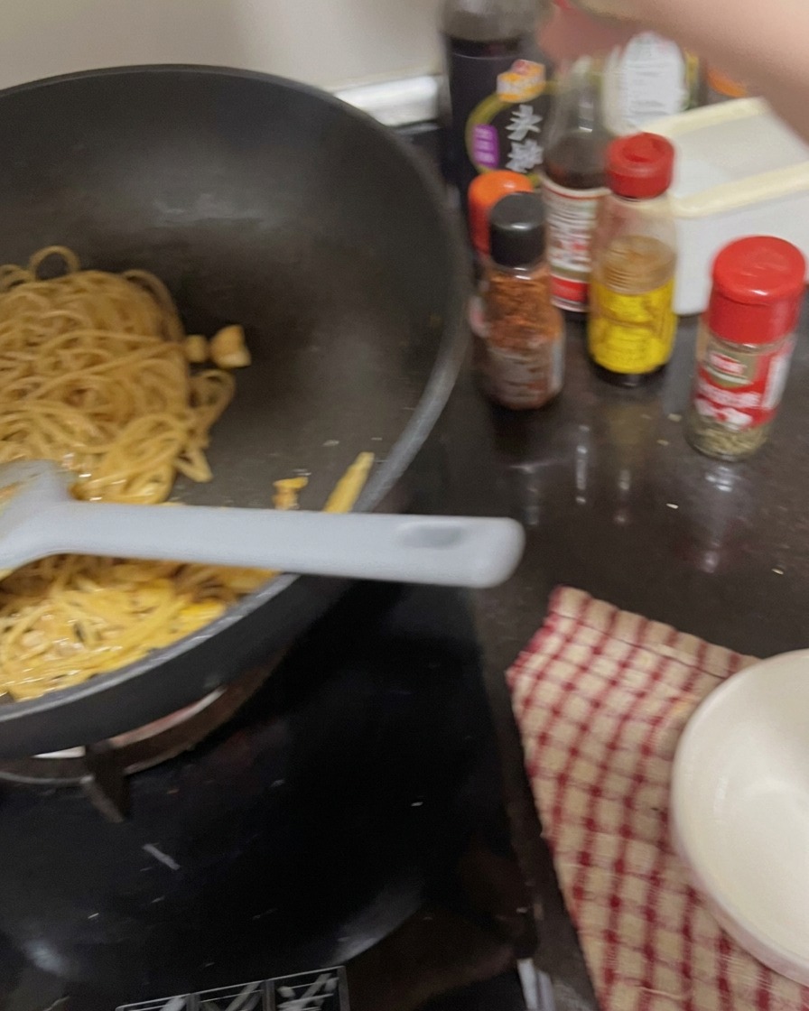 Eine Hand schüttelt einen Gewürzbehälter über der Pfanne mit Nudeln und Knoblauch.
