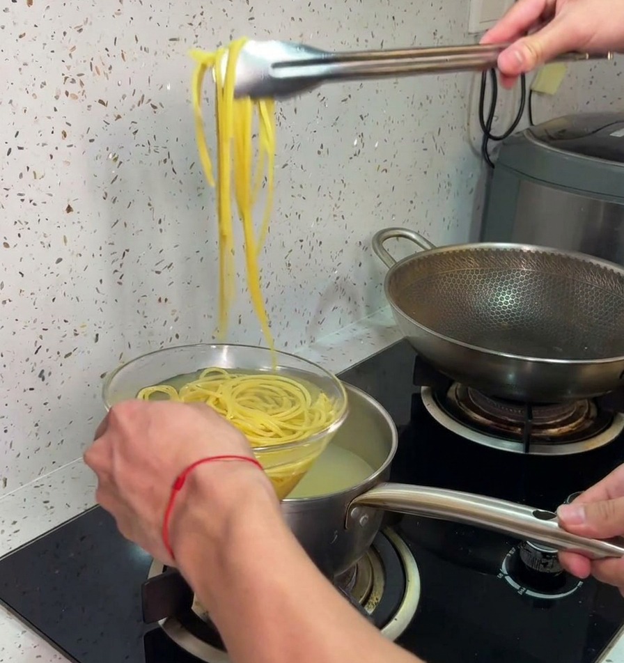 Eine Küchenzange hebt gekochte Spaghetti aus dem heißen Topf in eine Schüssel mit kaltem Wasser.