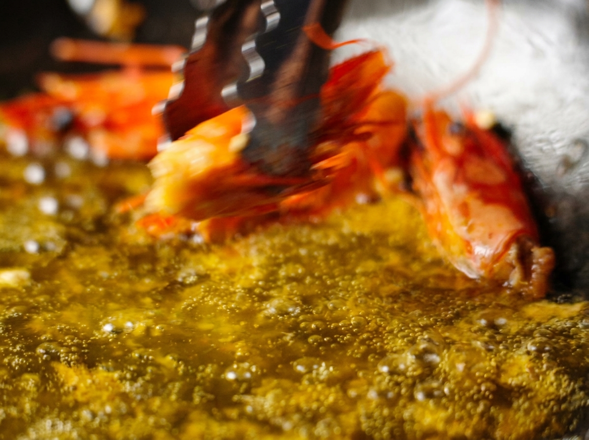 Eine Zange hebt einen frittierten Garnelenkopf aus einer Pfanne mit sprudelndem, orangefarbenem Öl.