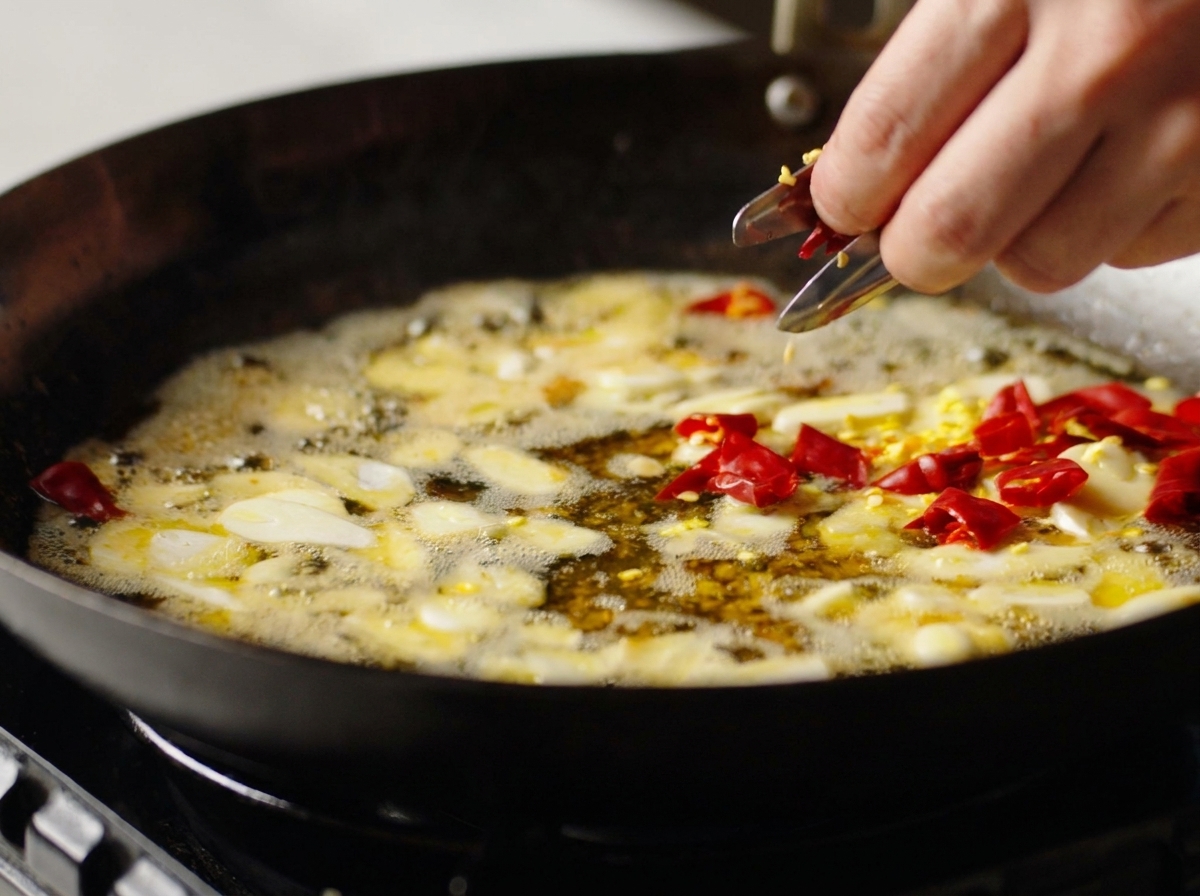 Eine Hand gibt getrocknete rote Chilistücke in einen Wok zu bratenem Knoblauch und gewürztem Öl.