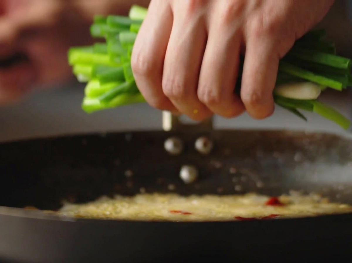 Eine Hand gibt einen großen Bund frisch gehackter Frühlingszwiebeln in einen heißen Wok mit sprudelndem Öl.