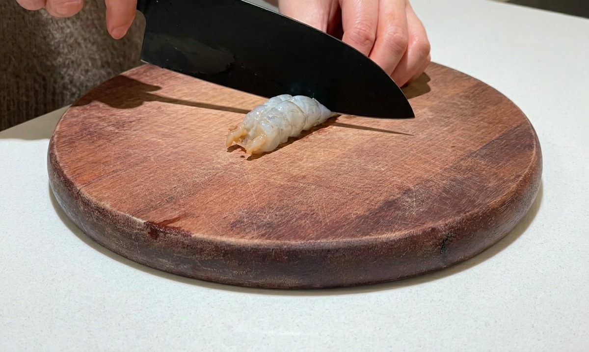Ein Messer schneidet entlang des Rückens einer geschälten rohen Garnele auf einem Holzbrett.