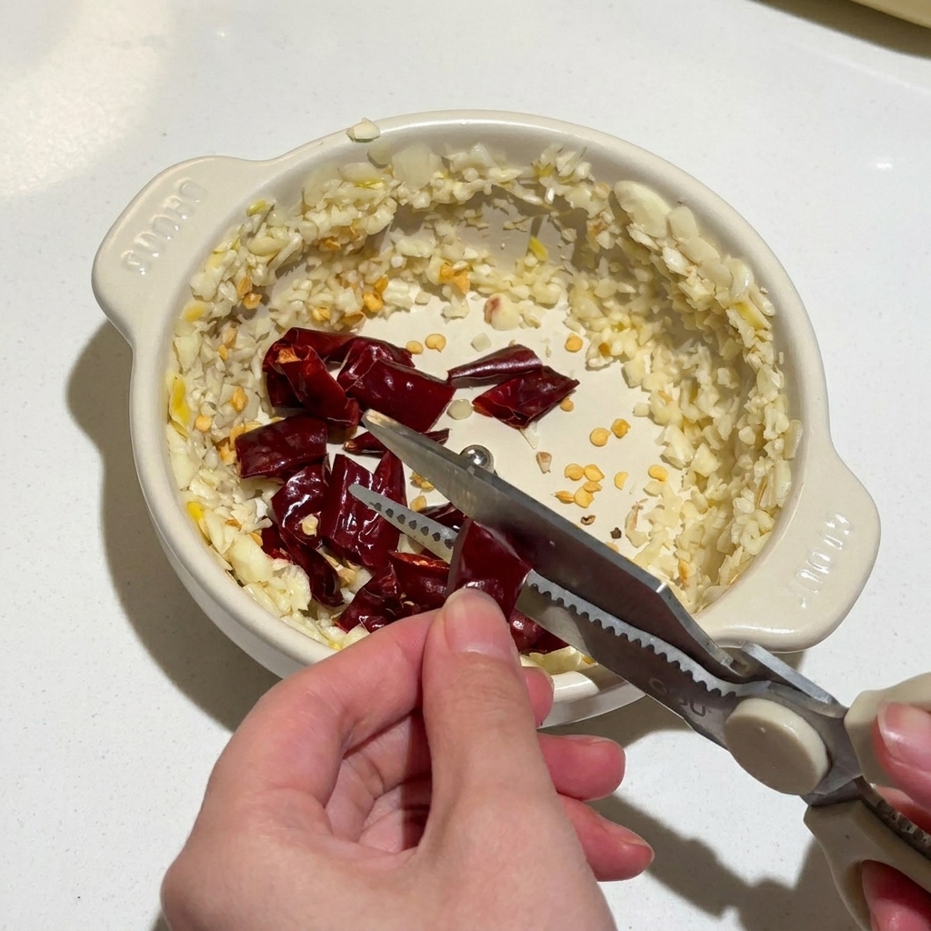 Hände schneiden mit einer Küchenschere getrocknete rote Chilischoten in eine Schüssel mit gehacktem Knoblauch.