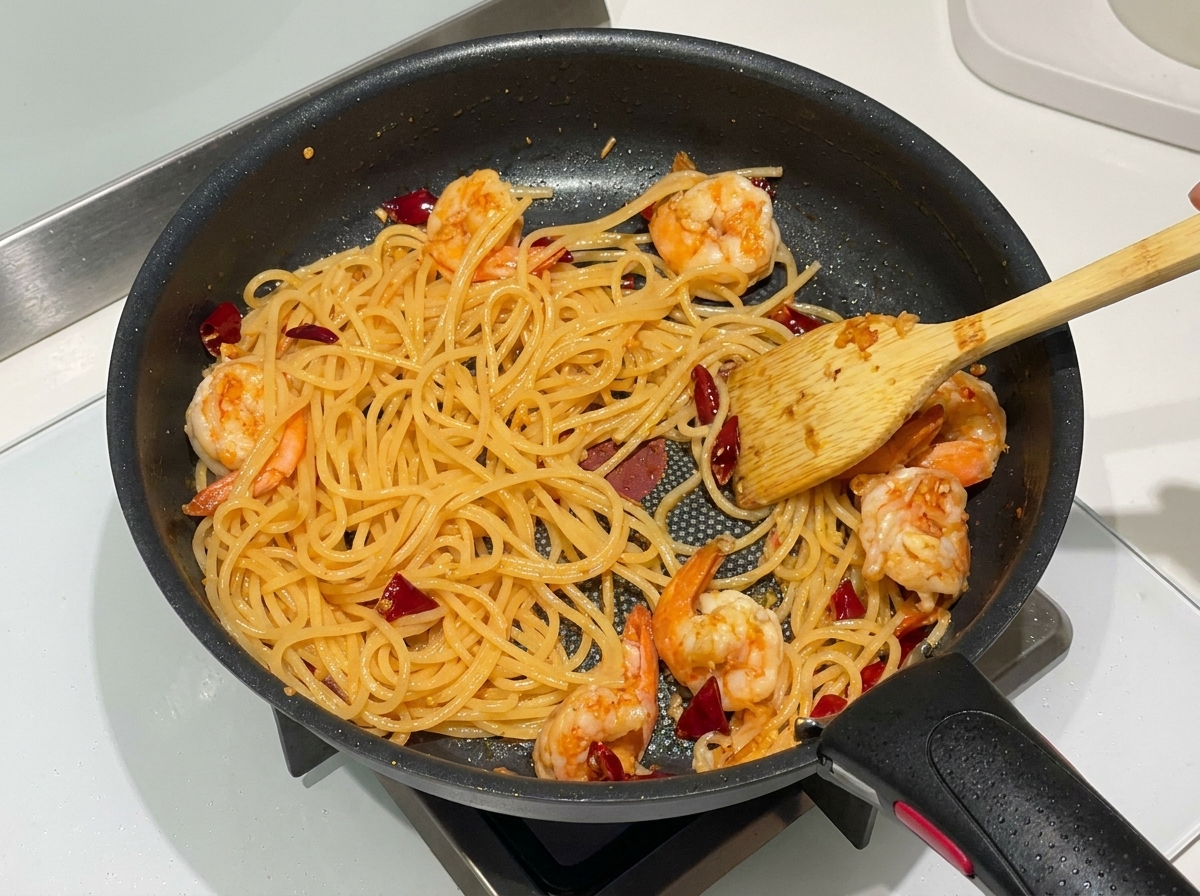 Gekochte Spaghetti, rosa Garnelen und getrocknete Chilis werden in einer Pfanne mit einem Holzspatel vermengt.