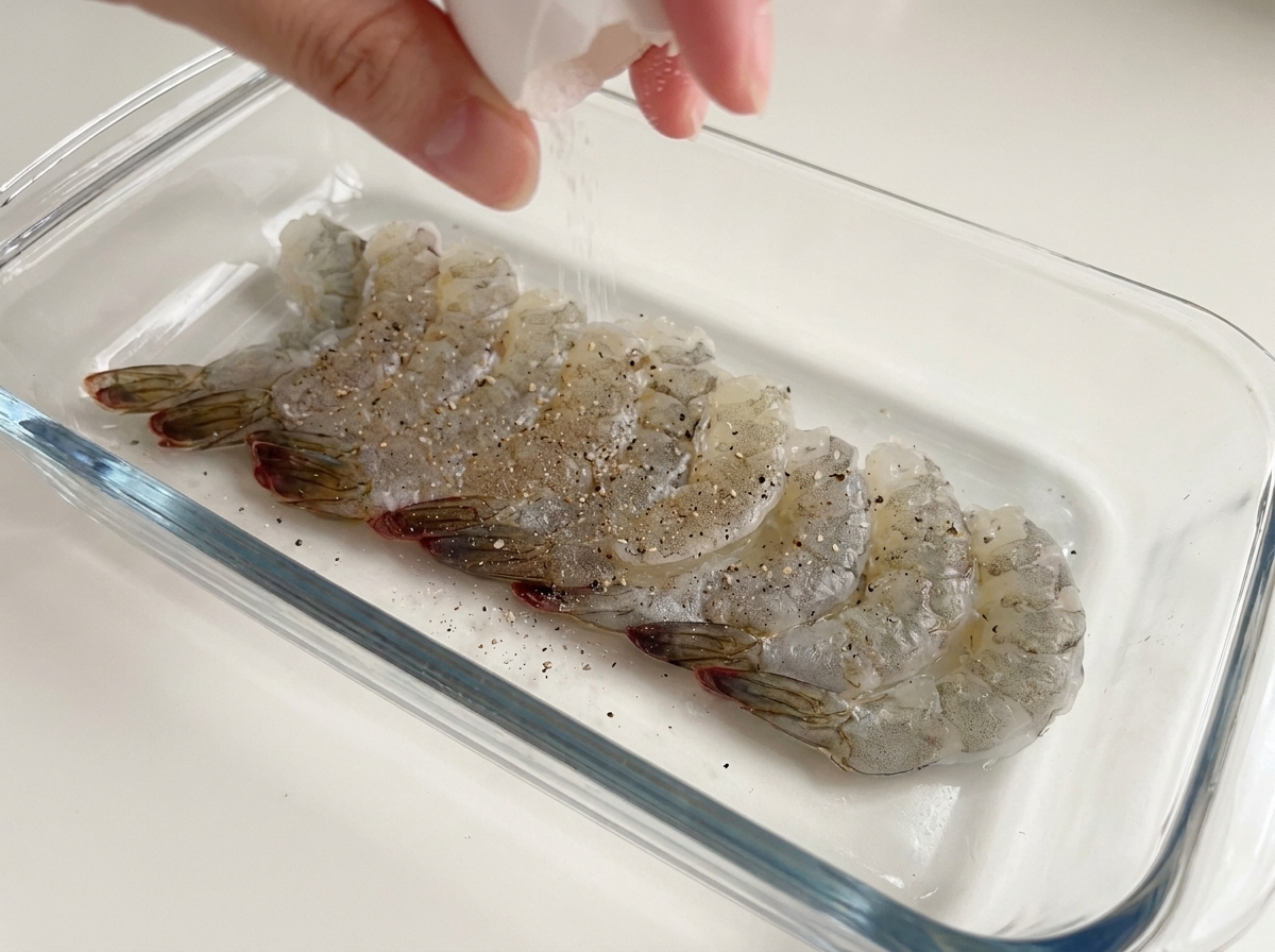 Eine Hand mahlt Meersalz und schwarzen Pfeffer über rohe Garnelen in einer Glasschale.