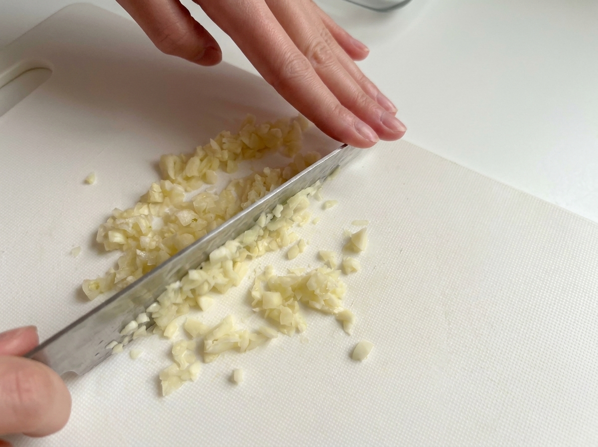 Hände hacken Knoblauchzehen mit einem Messer auf einem weißen Schneidebrett ganz fein.