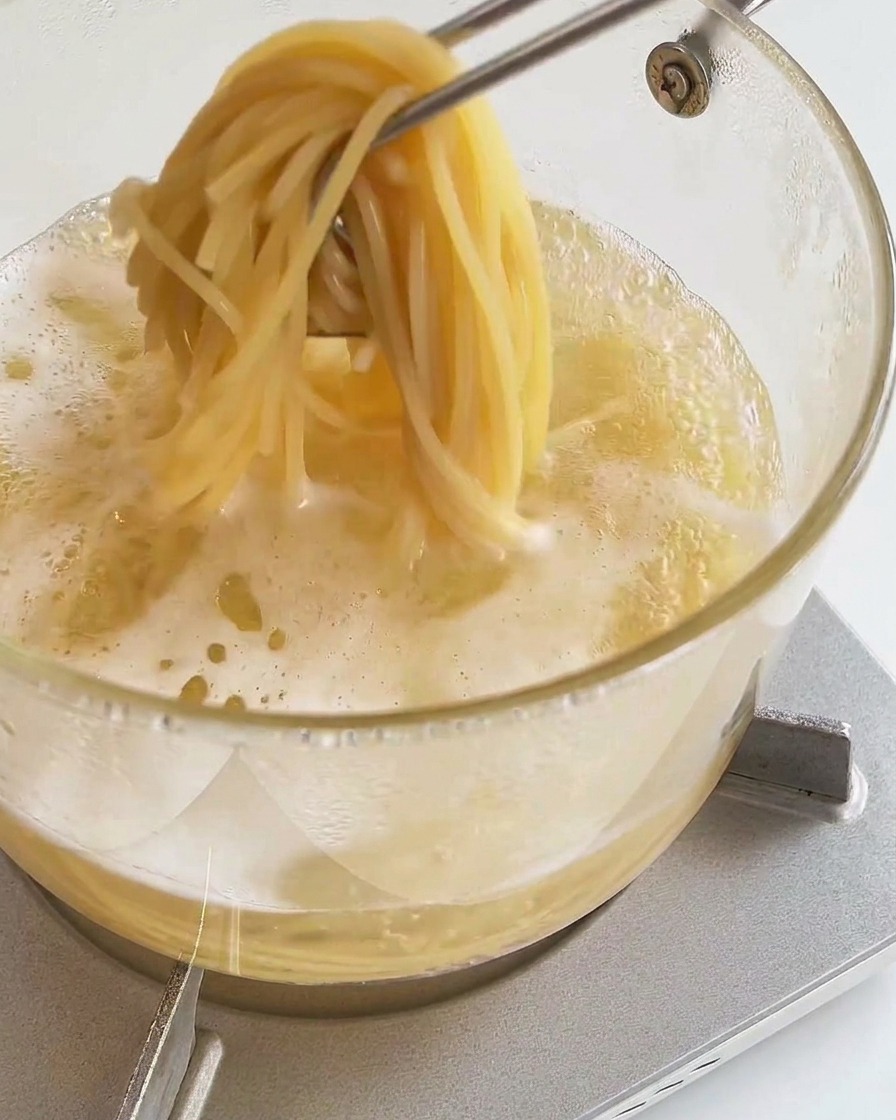 Eine Küchenzange hebt eine Portion gekochte Spaghetti aus dem dampfenden Topf.