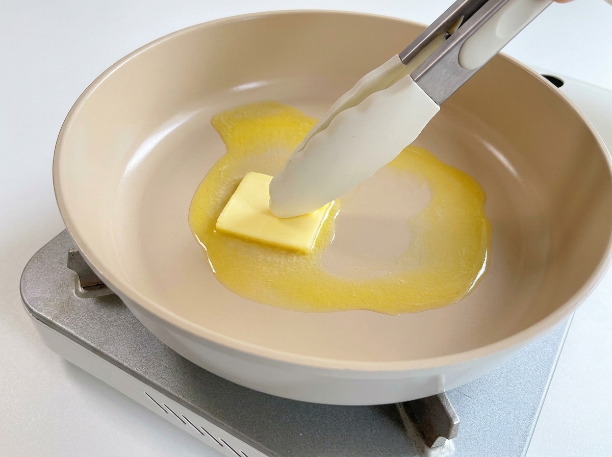 Ein Stück Butter schmilzt in einer heißen Pfanne und wird mit einer Zange bewegt.