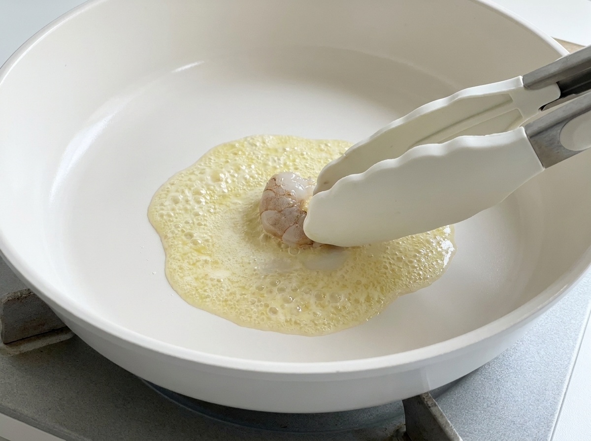 Eine Silikon-Zange legt eine rohe Garnele in die Pfanne mit schäumender Butter.