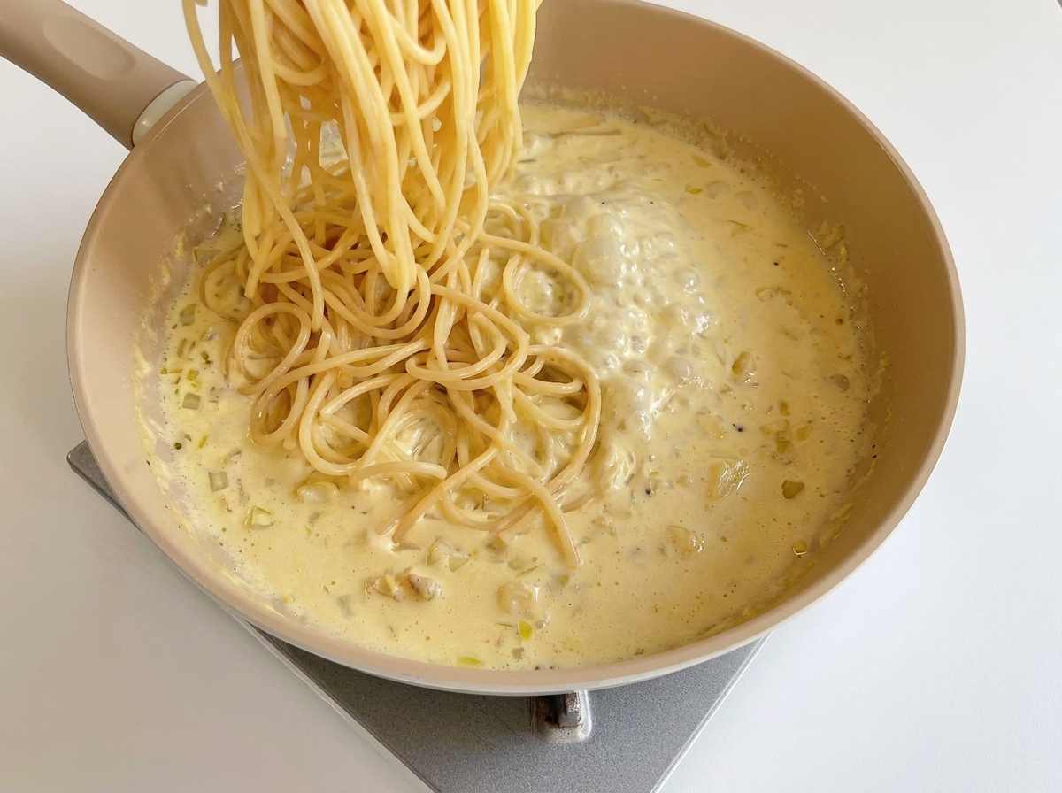 Gekochte Spaghetti werden in die Pfanne mit der cremigen Käsesauce gegeben.