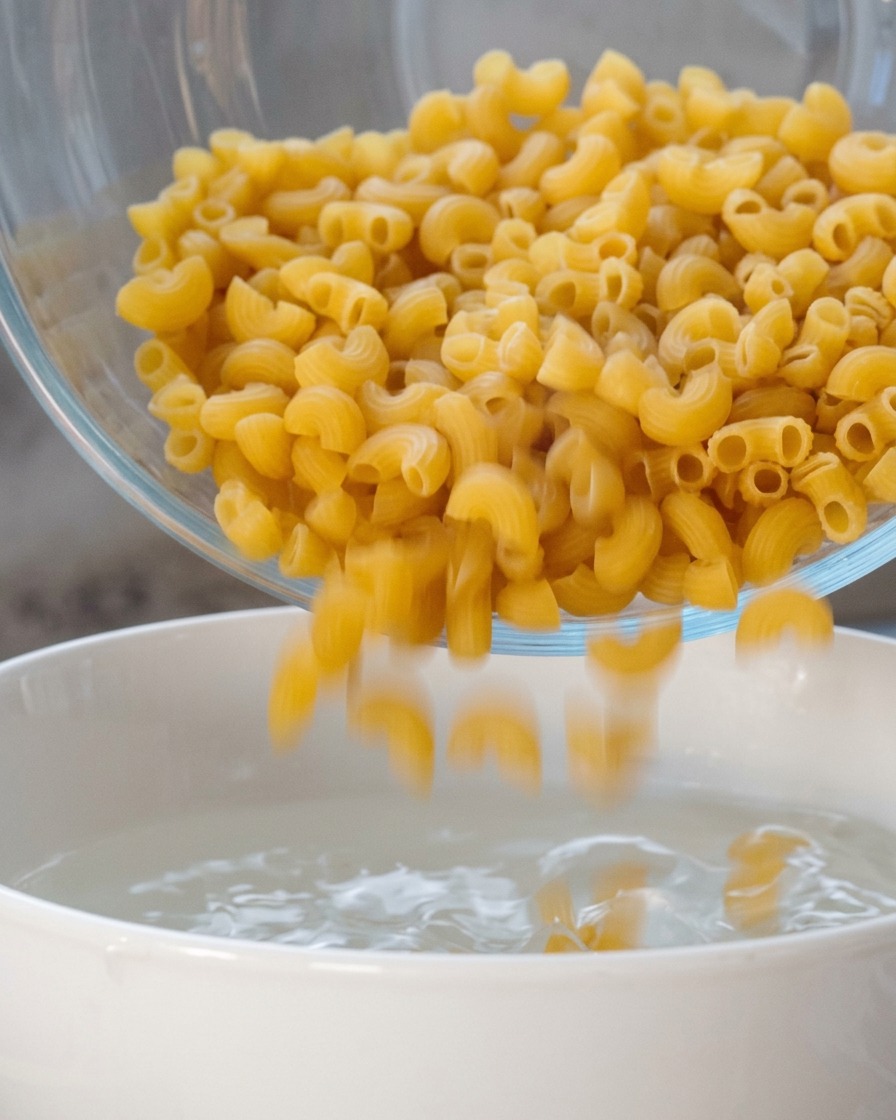 Trockene Spiralnudeln werden aus einer Glasschüssel in einen weißen Topf mit heißem Wasser gegossen.