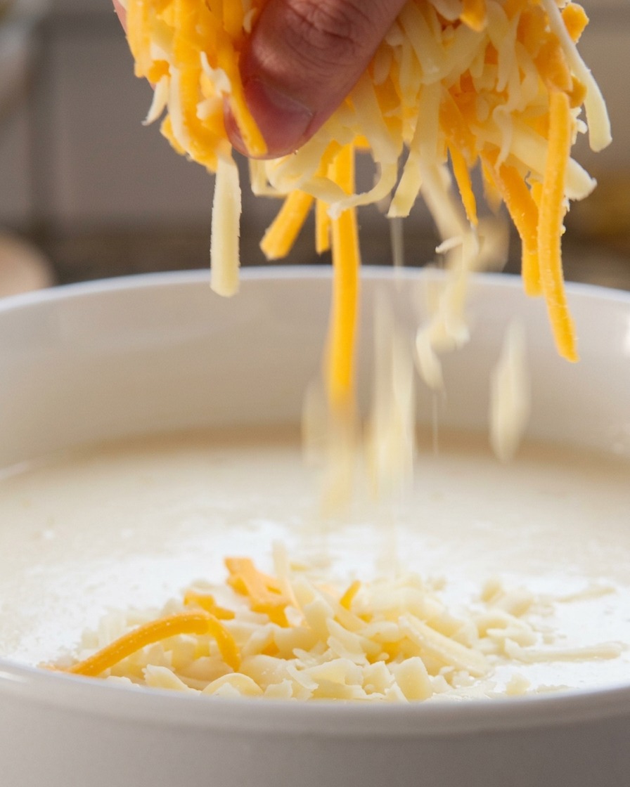 Eine Hand streut eine Mischung aus geriebenem weißem und orangem Käse in eine köchelnde weiße Flüssigkeit.
