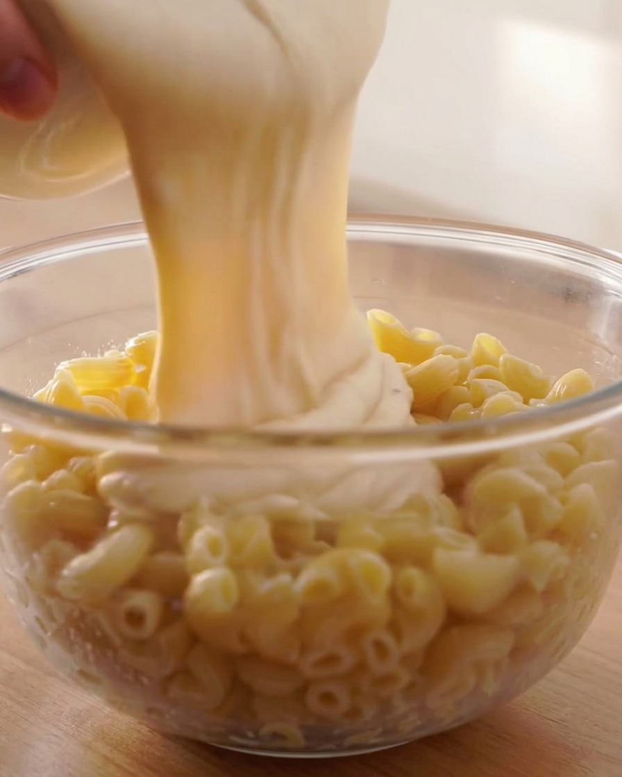 Dicke, cremige Käsesauce wird aus einem weißen Behälter über gekochte Spiralnudeln in einer Glasschüssel gegossen.