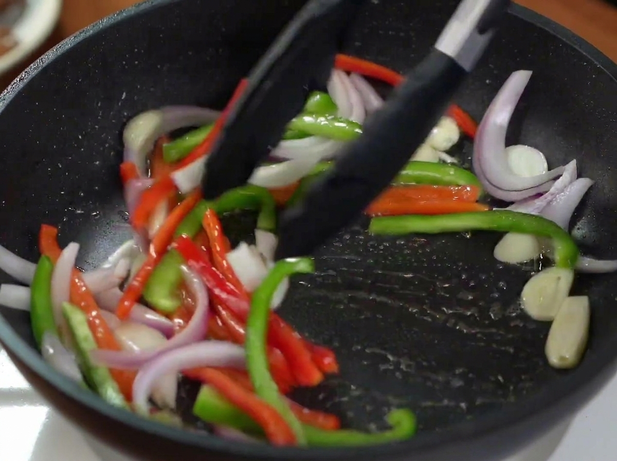Eine Grillzange wendet rote Zwiebeln und bunte Paprikastreifen in einer heißen Pfanne.