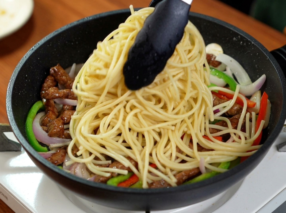 Gekochte Spaghetti werden mit einer Zange in die Pfanne zu Fleisch und Gemüse gegeben.