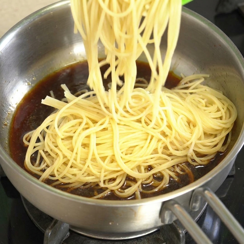 Gekochte Spaghetti werden in eine Edelstahlpfanne mit dunkler Rindfleisch-Pfeffer-Sauce gegeben.