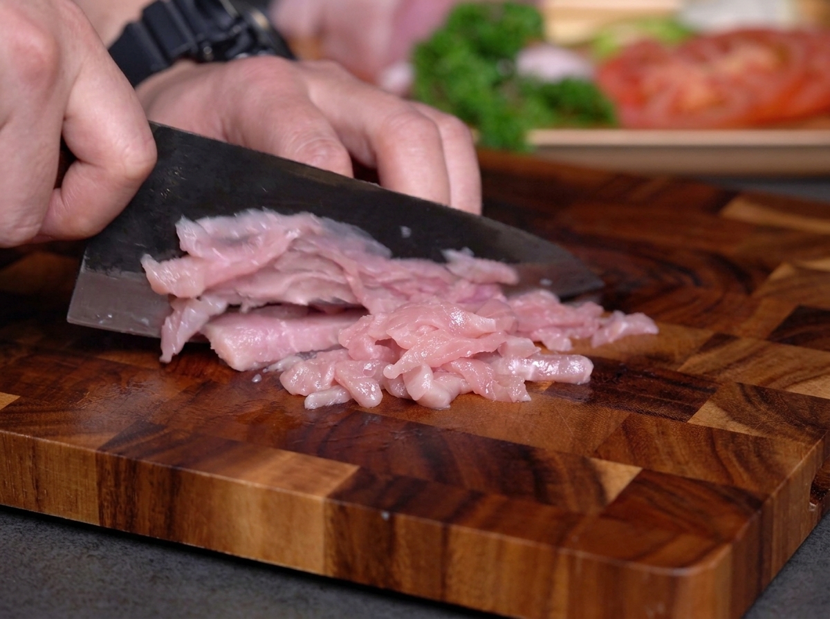 Hände schneiden rohes mageres Fleisch mit einem dunklen Messer auf einem Holzbrett in dünne Streifen.