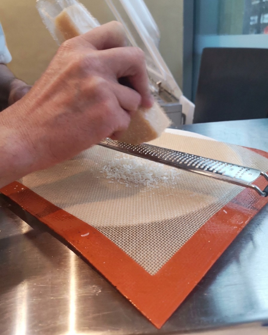 Hände reiben einen Block Parmesan mit einer feinen Microplane-Zesterreibe direkt auf eine Silikonbackmatte.