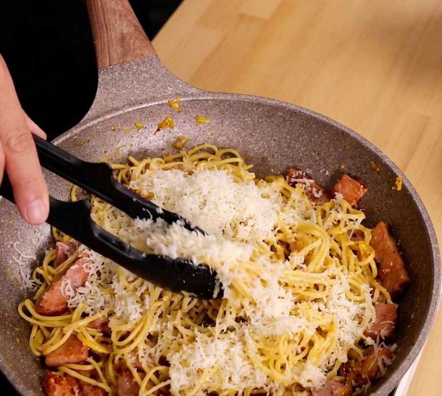 Hände vermengen Spaghetti, Käse und Speck mit einer Zange zu einer glänzenden Sauce.