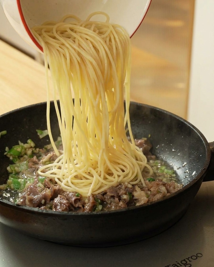 Gekochte Spaghetti werden aus einer Schüssel zum Fleisch in die Pfanne gegeben.