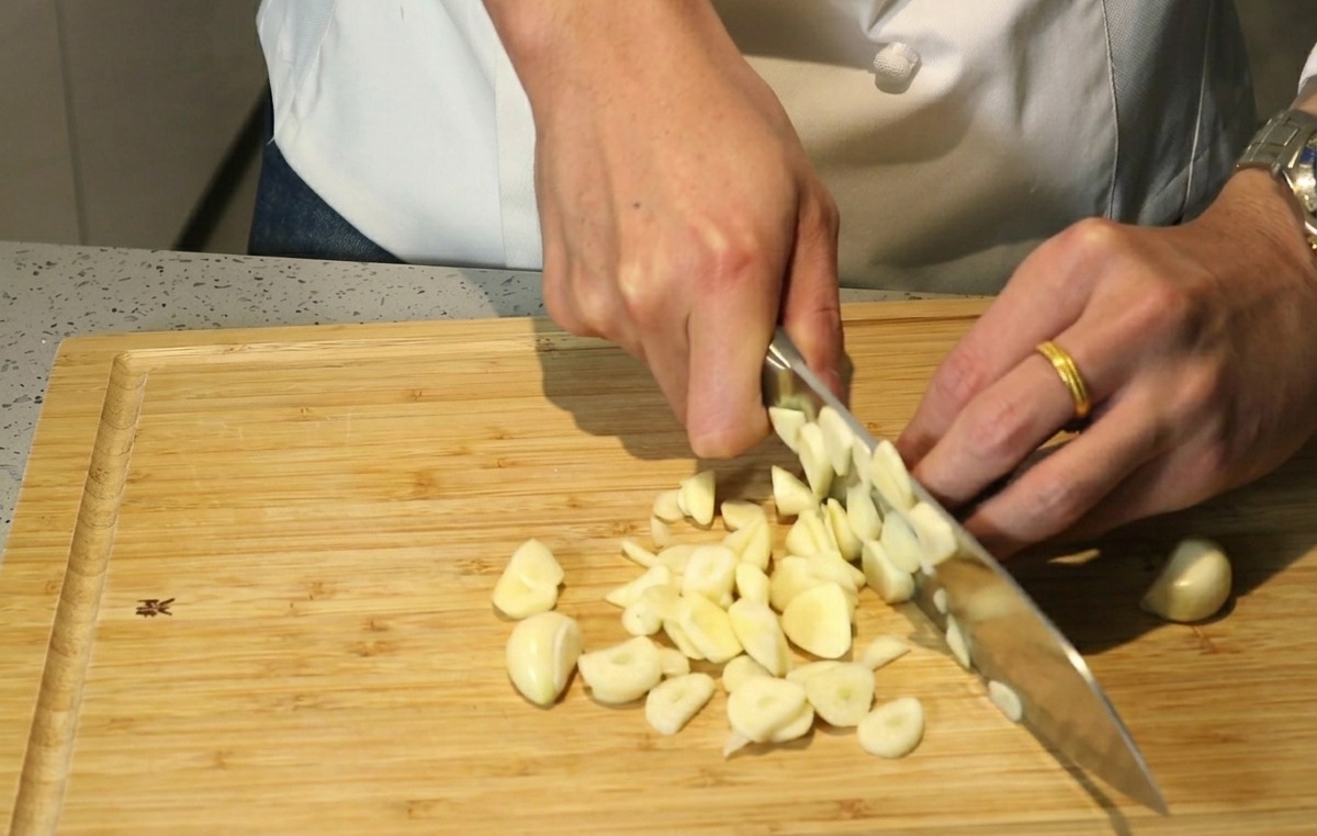 Hände schneiden Knoblauchzehen mit einem Messer auf einem Holzschneidebrett grob klein.