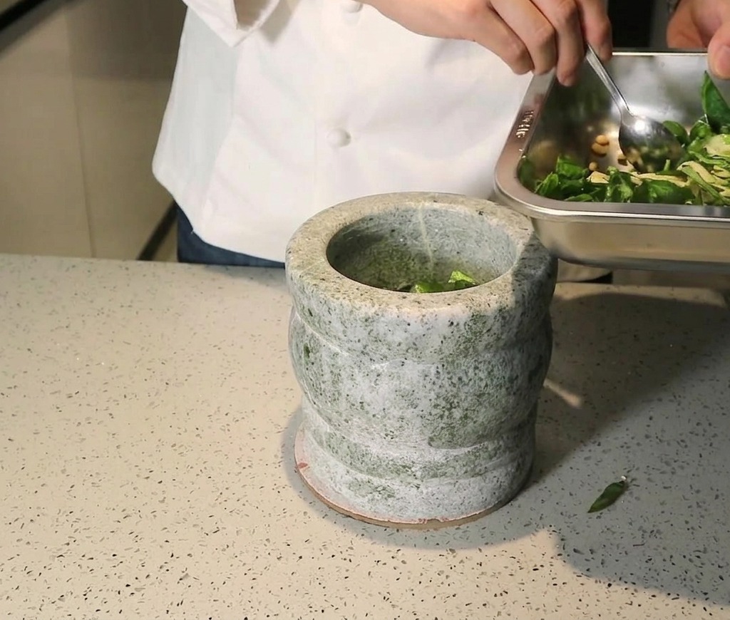 Basilikum, Pinienkerne und Knoblauch werden von einem Metallblech in einen grauen Steinmörser gegeben.