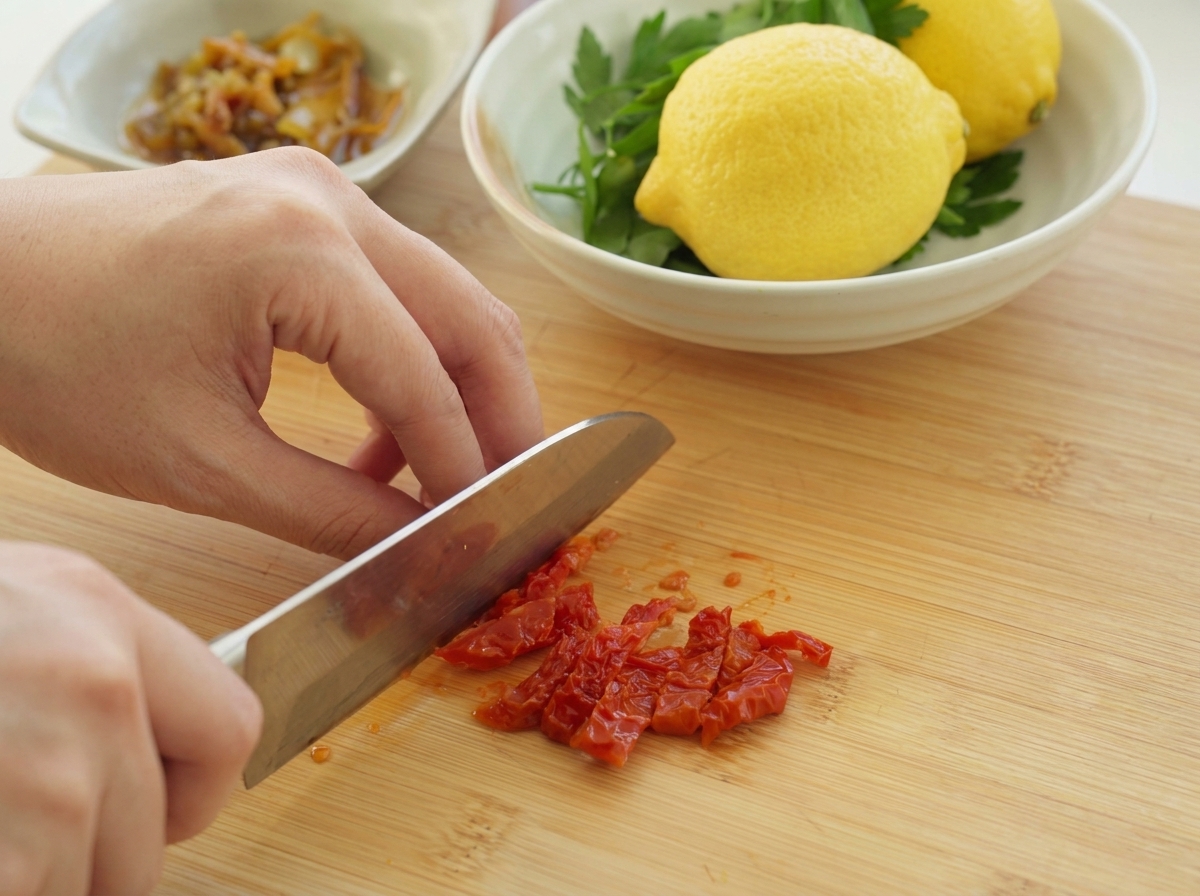 Eine Person schneidet leuchtend rote, getrocknete Tomaten mit einem Kochmesser auf einem Holzbrett.