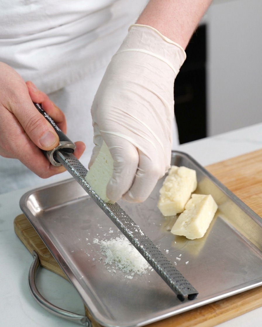 Hände reiben einen Block Pecorino-Käse mit einer Reibe fein auf eine Metallplatte.