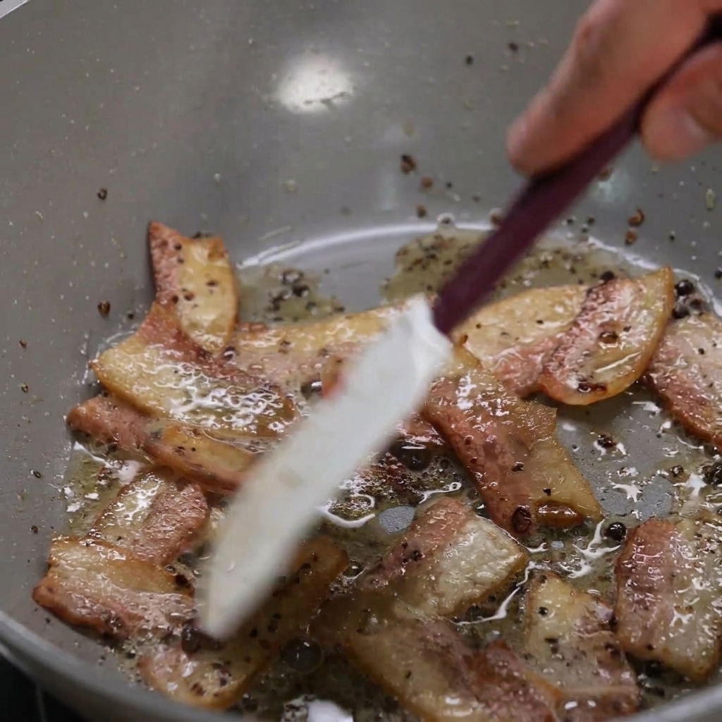 Guanciale-Streifen braten in einer Pfanne, während das Fett austritt und die Ränder knusprig werden.