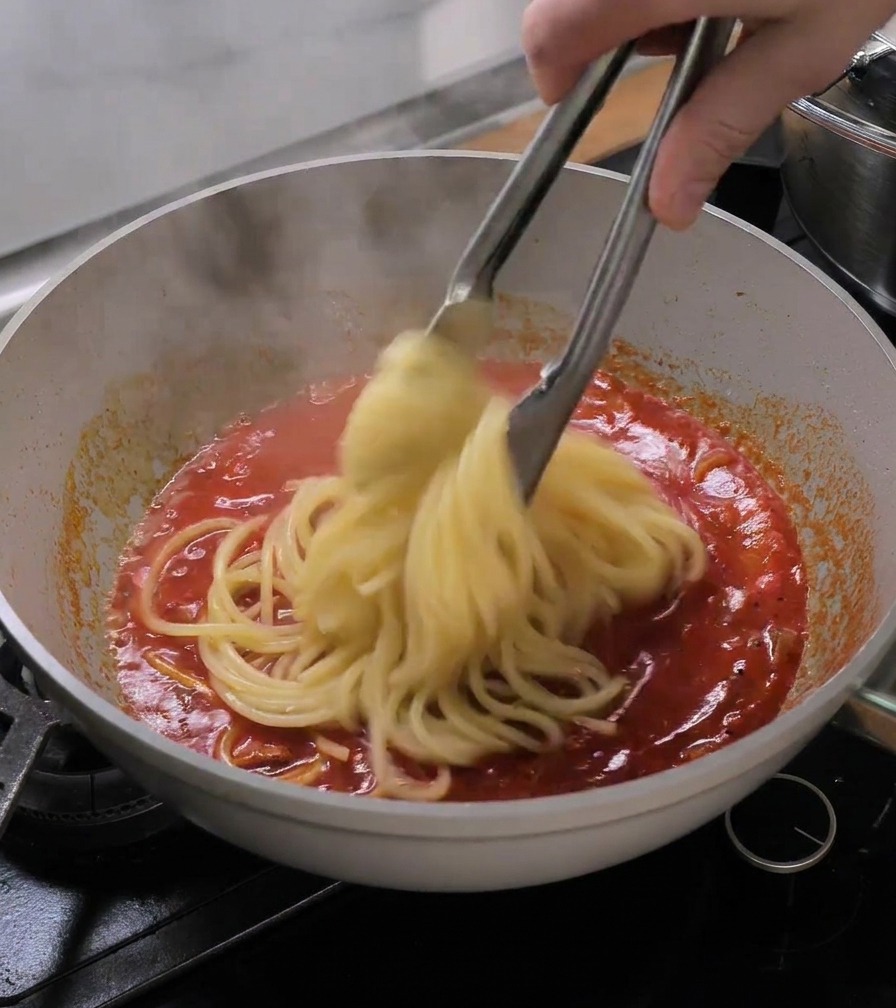 Eine Nudelzange hebt gekochte Bucatini direkt aus dem Wasser in die Pfanne mit der Tomatensauce.