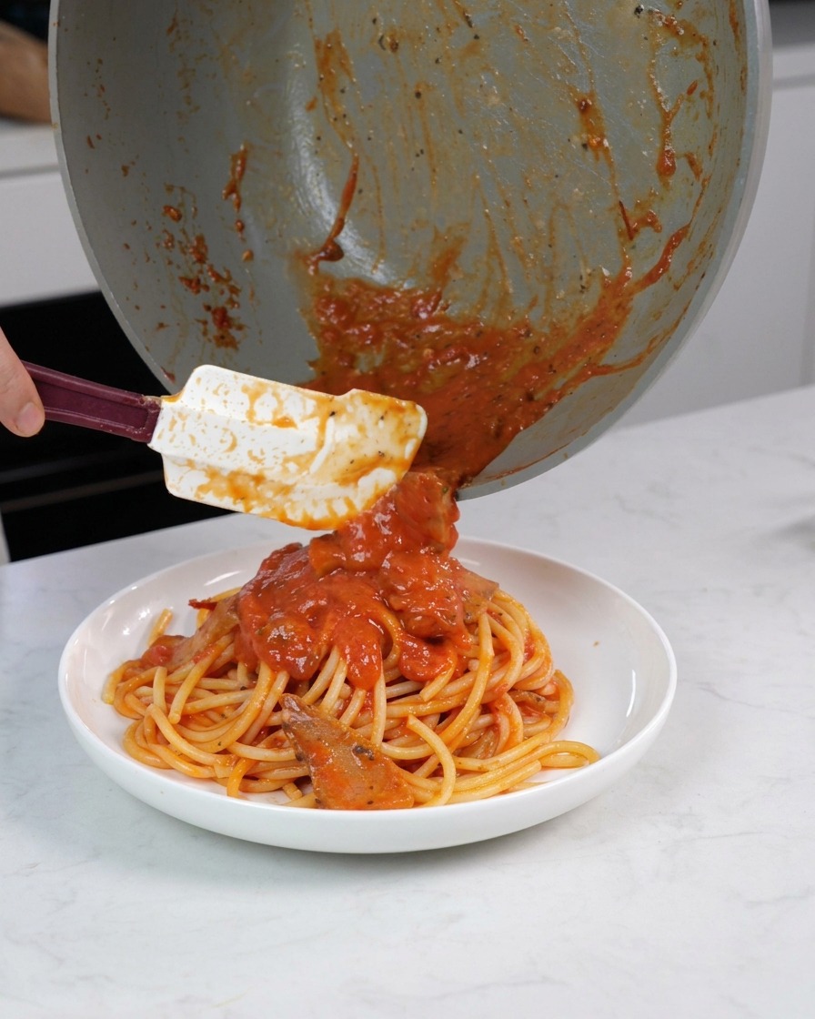 Ein Spatel schabt die restliche Sauce und Fleischstücke aus der Pfanne über die servierfertige Pasta im Teller.