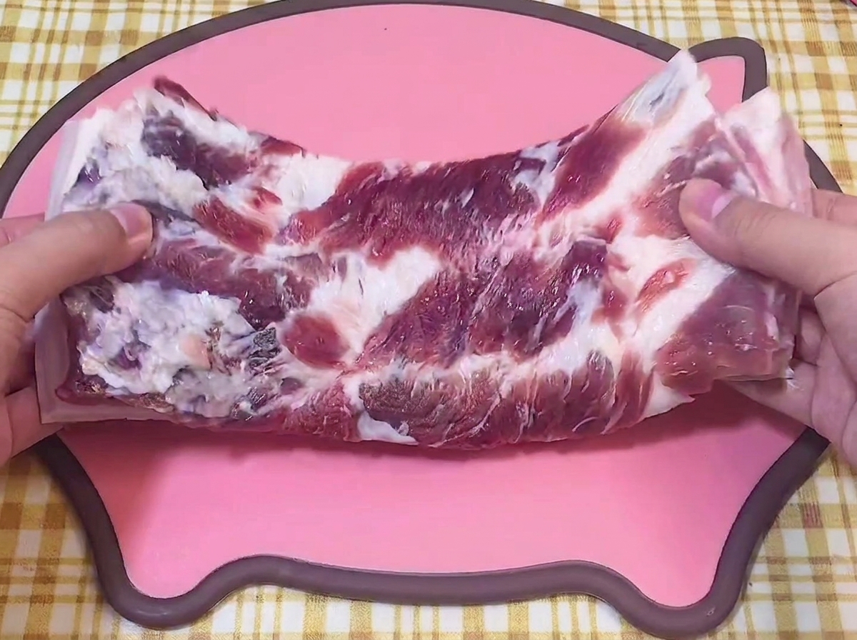 Hände halten ein Stück rohen Schweinebauch auf einem rosa Schneidebrett.