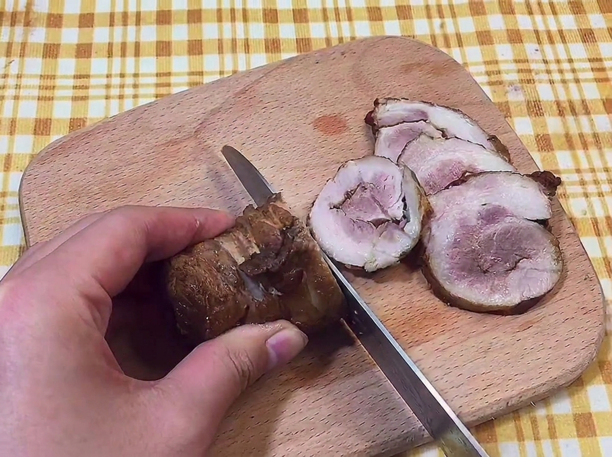 Ein Messer schneidet den Schweinebauch auf einem Holzbrett in dünne Scheiben.