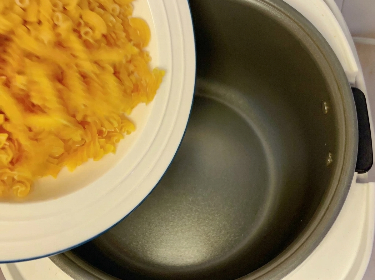 Trockene Fusilli-Nudeln werden von einem weißen Teller in den leeren Topf eines Reiskochers geschüttet.